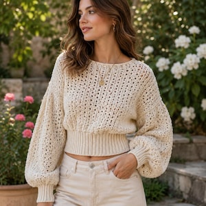 May include: A cream-colored, long-sleeved knit sweater featuring a wide, ribbed waistband and cuffs, paired with light-colored denim jeans. The sweater has a loose, open-weave texture throughout the bodice and sleeves, with a rounded neckline.