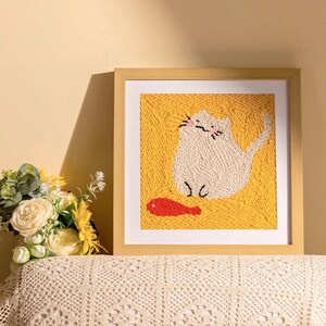 Puede incluir: Una obra de arte enmarcada que presenta un gato blanco y un pez rojo sobre un fondo amarillo. El gato está hecho de hilo texturizado, y el marco es de color marrón claro. La obra de arte se exhibe sobre una superficie blanca.