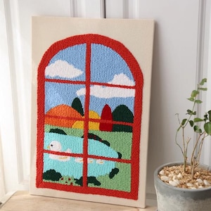 May include: A framed textile art piece featuring a window scene. The window frame is red, with a blue sky, white clouds, and a landscape of green, orange, and yellow. A light blue object is visible in the foreground.