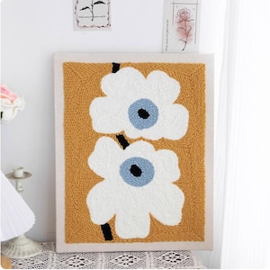 Puede incluir: Una obra de arte textil enmarcada con dos grandes flores blancas con centros azules y negros sobre un fondo amarillo mostaza. Las flores están conectadas por tallos negros. La obra está enmarcada en un borde crema.