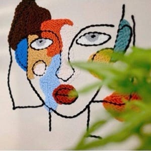 Op de afbeelding: Abstract geborduurde gezichtskunst met een modern ontwerp. Het kunstwerk toont een gezicht met kleurrijke secties in bruin, oranje, blauw en rood, omlijnd met zwarte draad. De ogen zijn grijs en de achtergrond is gebroken wit.