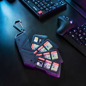 May include: A dark gray Switch game cartridge holder with a carabiner clip, holding several game cartridges. The holder is open, revealing the game cartridges with colorful game art. A keyboard and mouse are in the background.