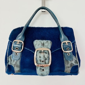 May include: A blue handbag with a textured fur-like finish, featuring teal snakeskin-patterned leather accents and three silver-toned buckles. The central buckle is embossed with the text DKNY, and the bag includes a single top handle.