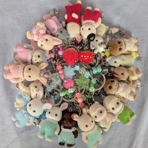 May include: A collection of flocked animal figurines attached to metal keychains, featuring various small creatures in colorful outfits, interspersed with decorative beads, star charms, and two distinct strawberry-shaped charms.