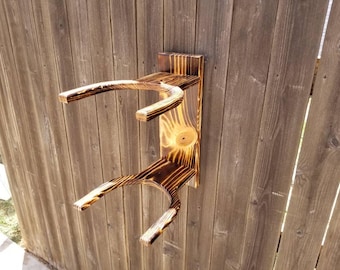 May include: A wall-mounted wooden rack featuring two U-shaped supports with a dark, scorched finish. The unit consists of a vertical rectangular backboard attached to two horizontal arms, showcasing natural wood grain and charred edges.