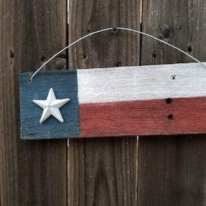 May include: A wooden sign painted with the Texas state flag. The sign is hanging on a wooden fence. The flag has a white five-pointed star in the blue section.