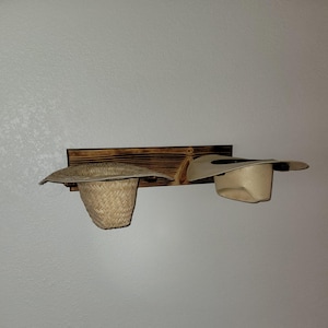May include: A rectangular, dark-stained wooden wall-mounted rack featuring two circular pegs. A woven straw hat with a wide brim hangs on the left peg, while a smooth, light-colored hat with a wide brim is positioned on the right peg.