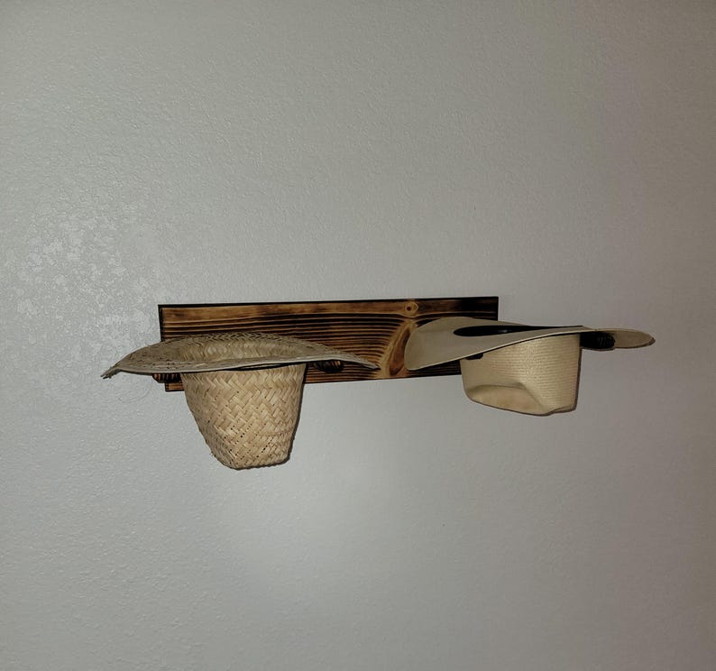 May include: A rectangular, dark-stained wooden wall-mounted rack featuring two circular pegs. A woven straw hat with a wide brim hangs on the left peg, while a smooth, light-colored hat with a wide brim is positioned on the right peg.