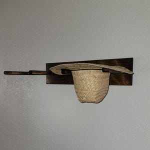 May include: A wall-mounted dark brown wooden rack featuring three horizontal pegs. A light tan woven straw hat is resting on the rightmost peg, with the brim extending over the wooden base and the crown hanging downward.