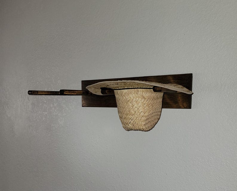 May include: A wall-mounted dark brown wooden rack featuring three horizontal pegs. A light tan woven straw hat is resting on the rightmost peg, with the brim extending over the wooden base and the crown hanging downward.