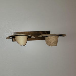 May include: A rectangular dark brown wooden wall-mounted rack featuring two horizontal supports. Two hats are resting on the rack: a smooth, light-colored straw hat on the left and a textured, woven straw hat on the right side.