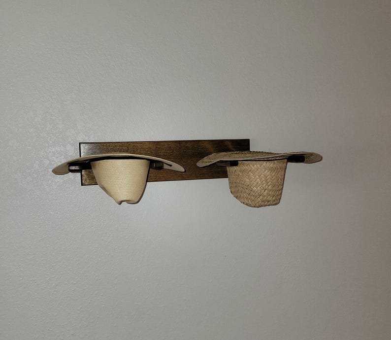 May include: A rectangular dark brown wooden wall-mounted rack featuring two horizontal supports. Two hats are resting on the rack: a smooth, light-colored straw hat on the left and a textured, woven straw hat on the right side.