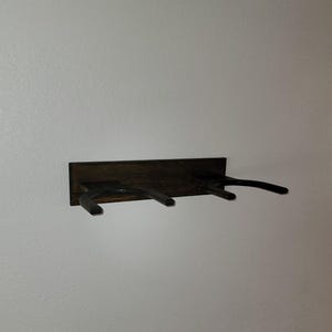 May include: A wall-mounted dark brown wooden rack featuring two horizontal U-shaped supports extending outward. The rectangular wooden base plate is mounted horizontally, providing a sturdy foundation for the two protruding arms designed to hold items.