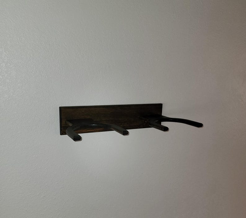 May include: A wall-mounted dark brown wooden rack featuring two horizontal U-shaped supports extending outward. The rectangular wooden base plate is mounted horizontally, providing a sturdy foundation for the two protruding arms designed to hold items.