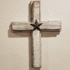 May include: A rustic wooden cross with a distressed white finish. A dark metal star is centered on the cross. The cross is made of reclaimed wood and is mounted on a beige wall.