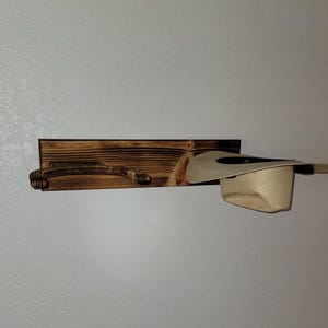 May include: A wall-mounted hat rack made of dark, scorched wood featuring a horizontal rectangular backboard and a curved support arm. A light beige cowboy hat is resting on the support arm, showcasing the functional design of the wooden rack.