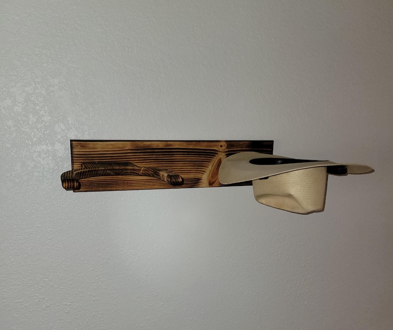 May include: A wall-mounted hat rack made of dark, scorched wood featuring a horizontal rectangular backboard and a curved support arm. A light beige cowboy hat is resting on the support arm, showcasing the functional design of the wooden rack.