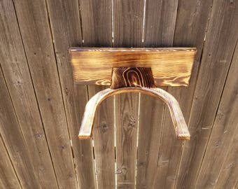 May include: A wooden shelf with a curved support, mounted on a wooden fence. The shelf has a rectangular top and a curved, arch-like support. The wood has a natural grain and a dark, burnt finish, creating a rustic aesthetic.