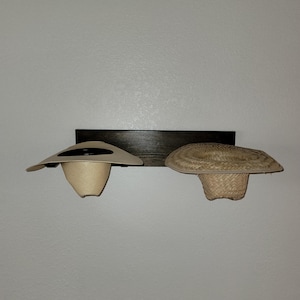 May include: A dark brown wooden wall-mounted rack featuring two hat holders. A light beige smooth straw hat is positioned on the left holder, while a textured, woven natural straw hat sits on the right holder, both displayed horizontally.