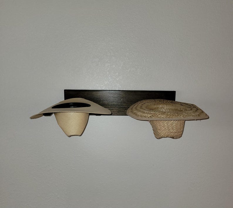 May include: A dark brown wooden wall-mounted rack featuring two hat holders. A light beige smooth straw hat is positioned on the left holder, while a textured, woven natural straw hat sits on the right holder, both displayed horizontally.