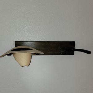 May include: A dark brown wooden wall-mounted rack featuring two horizontal pegs. A light beige straw hat with a dark band is resting on the left peg, while the right peg remains empty and extends outward from the rectangular wooden base.