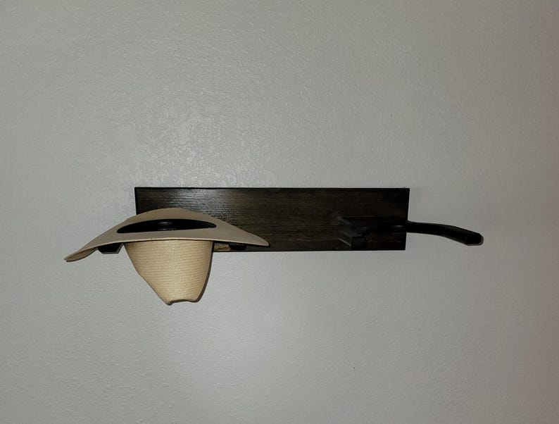 May include: A dark brown wooden wall-mounted rack featuring two horizontal pegs. A light beige straw hat with a dark band is resting on the left peg, while the right peg remains empty and extends outward from the rectangular wooden base.