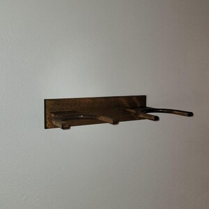 May include: A dark brown stained wooden wall mount rack featuring two U-shaped horizontal supports extending outward from a rectangular backplate. The wood grain is visible throughout the structure, which is designed to hold two items.