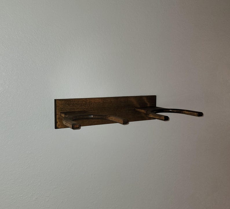 May include: A dark brown stained wooden wall mount rack featuring two U-shaped horizontal supports extending outward from a rectangular backplate. The wood grain is visible throughout the structure, which is designed to hold two items.