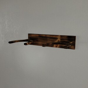 May include: A wall-mounted wooden rack featuring a dark, scorched finish with two U-shaped supports extending outward. The rectangular backboard displays a prominent wood grain pattern with charred edges and a deep brown, burnt aesthetic.