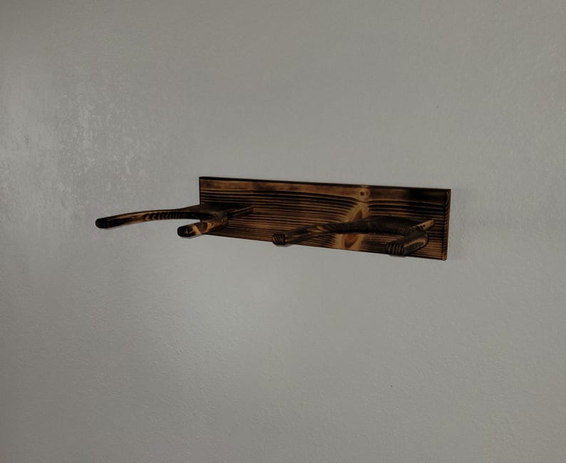 May include: A wall-mounted wooden rack featuring a dark, scorched finish with two U-shaped supports extending outward. The rectangular backboard displays a prominent wood grain pattern with charred edges and a deep brown, burnt aesthetic.