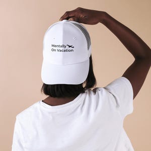 以下が含まれることがあります： メッシュバックとカーブしたつばが付いた白いトラッカーハット。「Mentally On Vacation」の文字と小さな飛行機のグラフィックが黒でプリントされています。帽子は人が持っています。
