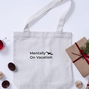 以下が含まれることがあります： 「Mentally On Vacation」の文字と飛行機のイラストがプリントされた白いトートバッグ。長い持ち手があり、プレゼントや紙製のオーナメントなど、装飾的なアイテムに囲まれています。