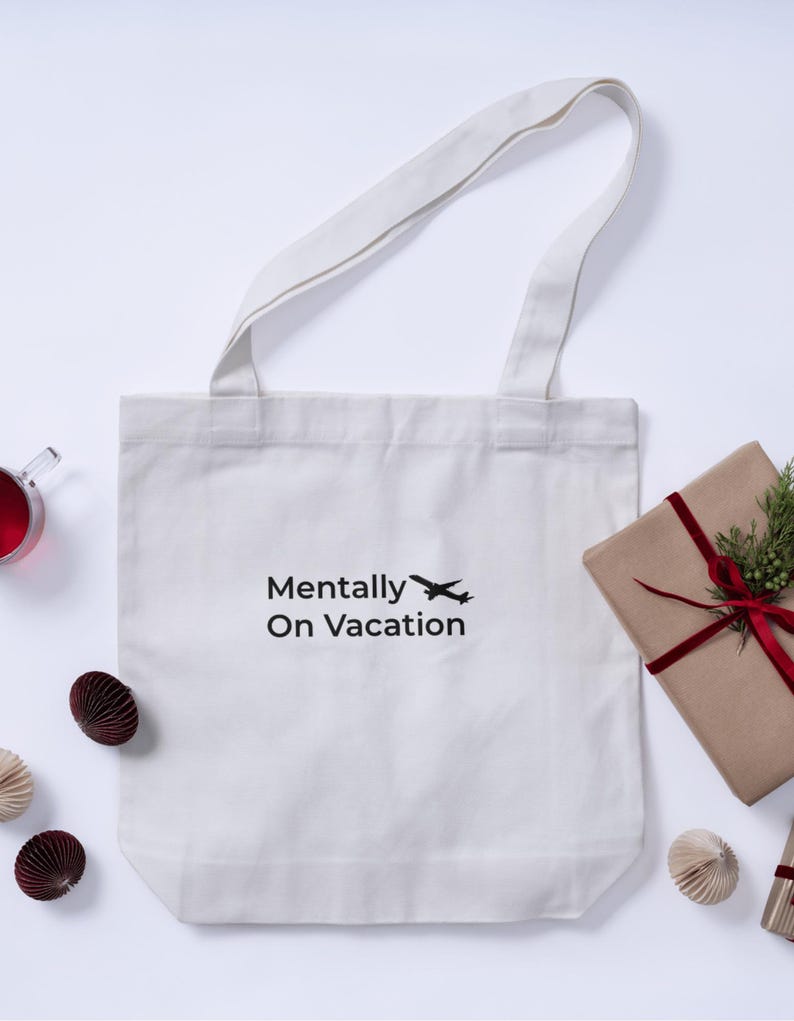 以下が含まれることがあります： 「Mentally On Vacation」の文字と飛行機のイラストがプリントされた白いトートバッグ。長い持ち手があり、プレゼントや紙製のオーナメントなど、装飾的なアイテムに囲まれています。