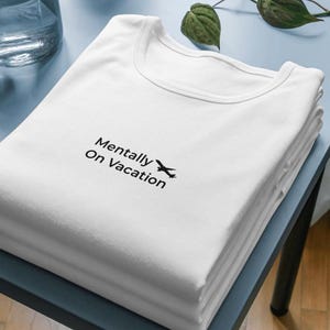 以下が含まれることがあります： 「Mentally On Vacation」の文字と小さな飛行機のグラフィックが黒でプリントされた白いTシャツの山。シャツは水色の表面にきれいに折りたたまれています。