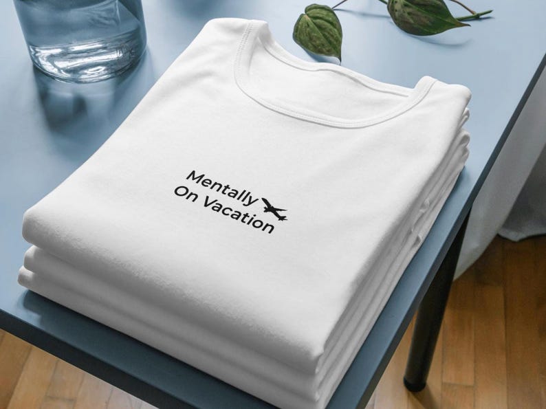 以下が含まれることがあります： 「Mentally On Vacation」の文字と小さな飛行機のグラフィックが黒でプリントされた白いTシャツの山。シャツは水色の表面にきれいに折りたたまれています。