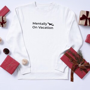 以下が含まれることがあります： 「Mentally On Vacation」の黒い文字と小さな飛行機のグラフィックがプリントされた白いスウェットシャツ。赤と金のギフトと装飾的な要素が配置されています。