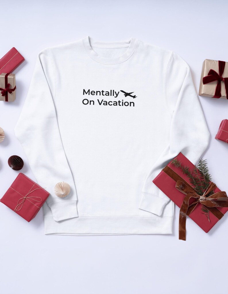 以下が含まれることがあります： 「Mentally On Vacation」の黒い文字と小さな飛行機のグラフィックがプリントされた白いスウェットシャツ。赤と金のギフトと装飾的な要素が配置されています。