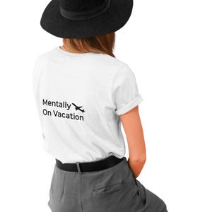 以下が含まれることがあります： 黒い帽子、白いTシャツ、グレーのズボンを着用した人。白いTシャツには「Mentally On Vacation」の文字と小さな飛行機のグラフィックが入っています。その人はスツールに座っています。