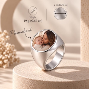 Puede incluir: Un anillo de sello plateado con una inserción circular que muestra una fotografía en color de una persona besando a un bebé. El anillo pesa 19 g y tiene un diámetro de 2 cm. La palabra Personalized está impresa junto al anillo.