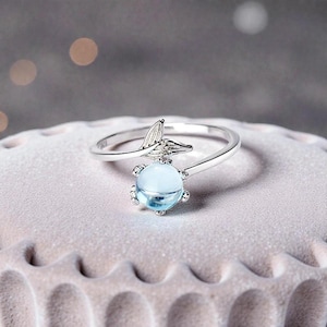 May include: A silver ring with a light blue, round gemstone. The ring has a tail-like design above the gemstone. The ring is displayed on a light pink, textured surface.
