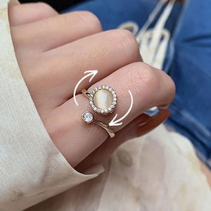 May include: Gold-toned adjustable ring featuring a central, round, iridescent stone surrounded by a halo of small, clear crystals. A smaller, clear crystal accents the band. The ring is designed with a rotating feature, indicated by white arrows.