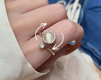 May include: Gold-coloured adjustable ring with a central, round, iridescent stone surrounded by small, clear crystals. A smaller, clear crystal accents the band. The ring has a rotating feature, indicated by white arrows.