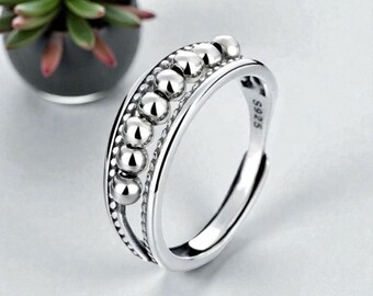 May include: A silver fidget ring featuring a row of small, spherical beads. The ring has a polished finish and is adjustable. The background includes a small potted plant.