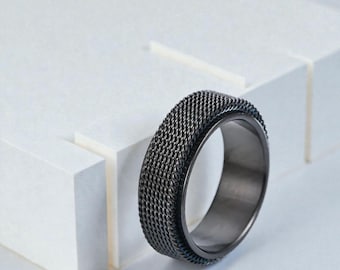 May include: A dark grey ring featuring a woven mesh band. The ring has a smooth inner surface and a textured outer surface. The ring is set against a white background with geometric shapes.