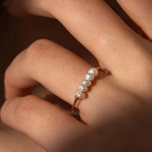 Può includere: Un delicato anello d'oro con una fila di piccole perle bianche dalla forma irregolare. La fascia dell'anello è sottile e dorata, e le perle sono raggruppate. L'anello è indossato su un dito, mettendo in risalto l'elegante design del gioiello.