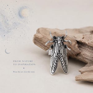 May include: A silver-toned ring featuring a detailed cicada insect design with textured wings and legs. The ring is displayed alongside text that reads: FROM NATURE TO INSPIRATION. Wear the art. Live the story.