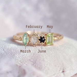 Puede incluir: Un anillo de oro con cuatro piedras preciosas de diferentes formas y colores: una marquesa verde, un óvalo transparente, una piedra negra en forma de flor y una esmeralda verde tallada. El anillo está etiquetado con los meses febrero, marzo, mayo y junio.