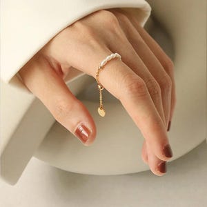 May include: A delicate gold ring featuring a string of small pearls on the finger. A fine gold chain extends from the ring, culminating in a small heart-shaped charm. The ring is worn on a hand with polished nails.