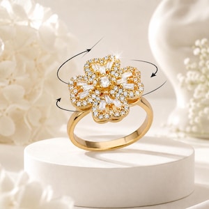 May include: A gold-toned ring featuring a four-leaf clover design. The clover is encrusted with numerous small, clear, round-cut stones and several baguette-cut stones, with a single round-cut stone at the centre. Curved arrows indicate rotation.