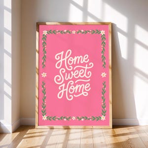 May include: A framed art print with a pink background and the words "Home Sweet Home" in white script. The print is bordered by a green and white floral design and encased in a wooden frame. The artwork is leaning against a wall.